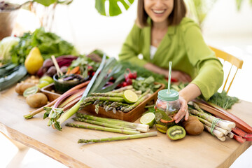 Young woman takes a bottle with drink while sitting by the table full of fresh vegetables, fruits and greens indoors. Concept of healthy vegan food and lifestyle