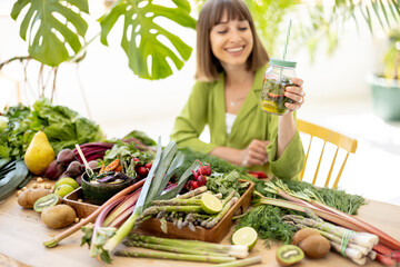 Obraz premium Young woman takes a bottle with drink while sitting by the table full of fresh vegetables, fruits and greens indoors. Concept of healthy vegan food and lifestyle