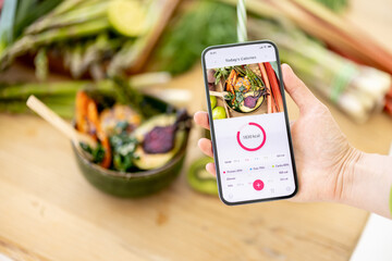 Holding smart phone with running mobile application for track calories consumed on background of healthy food ingredients. Concept of modern technology in proper nutrition - obrazy, fototapety, plakaty