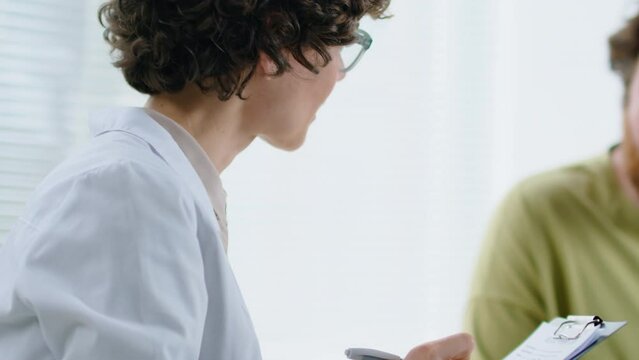 Tilt up shot of female doctor listening to male patient and taking notes on clipboard during medical consultation in clinic