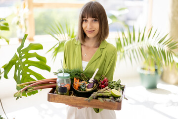 Portrait of cheerful young woman with tray full of fresh food ingredients and vegetarian lunch in...
