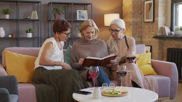 Three modern mature women looking at photos in photo album and remembering good times, sitting on sofa in loft style living room drinking red wine and chatting