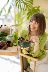 Young cheerful woman eats vegetarian lunch in bowl, sitting by the table full of fresh food...