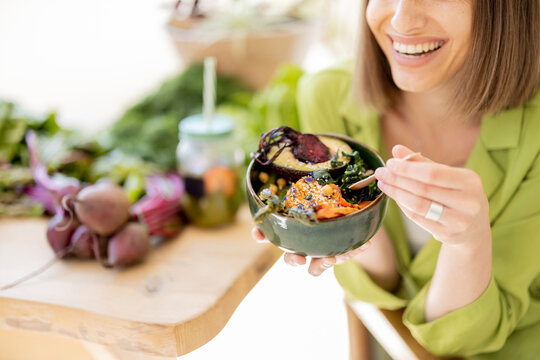Young cheerful woman eats vegetarian lunch in bowl, sitting by the table full of fresh food ingredients indoors, close-up. Healthy lifestyle and wellness concept
