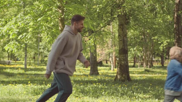 PAN Slowmo Of Caucasian Man And His 7 Year Old Son Having Fun Outdoors In Park On Sunny Day, Playing Catch Up Game Around Tree