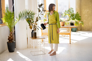 Young woman in apron as housewife or gardener takes care of green plants, watering flower pots in...