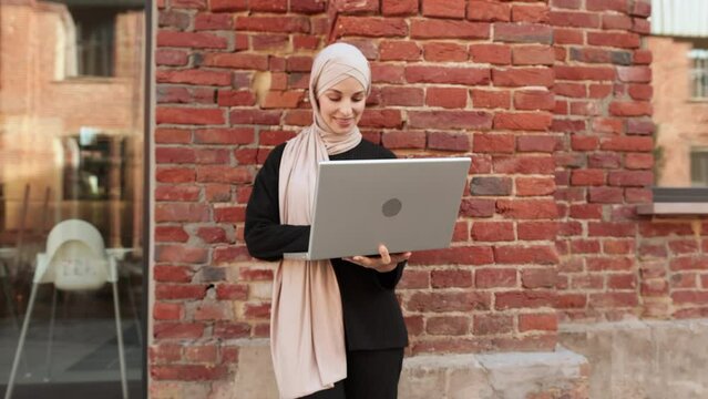 Cheerful young islamic businesswoman working on laptop while standing near her office. Beautiful young woman with hijab working using laptop during break.