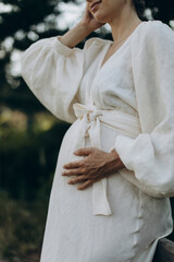 a young happy woman in a white voluminous linen dress holds her hand on her pregnant tummy. body of pregnant woman in white dress closeup