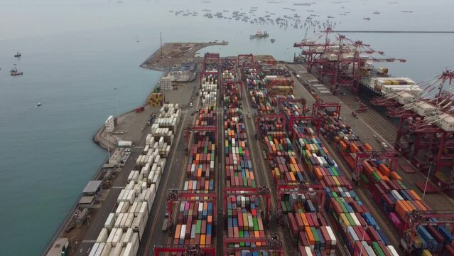 Drone video of shipping container yard in sea port of Callao in Peru. Many colored containers can be seen and ships off in the horizon. Recorded on a cloudy morning. Top down, tilting camera up.