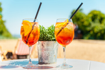 Two glasses of orange spritz aperol drink cocktail on table outdoors with sea and trees view blurred background.