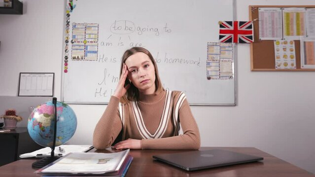 Upset female english teacher sitting at school desk with laptop and looking at camera. Tired woman leaning head on hand and getting lost in sad thoughts about work.