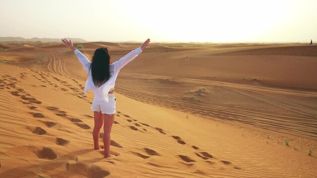 Young woman tourist in Dubai rear view from back stands in desert and stretches arms forward and sunbathes under sunlight enjoying warm weather. Traveling on vacation to beautiful places of world.
