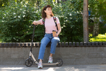 middle aged woman with a backpack and scooter rests in the park and drinks coffee on a summer day