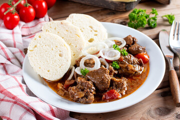 Pork goulash meat with dumplings on white plate, cutlery, garlic, onion, tablecloth in the...