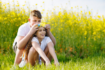 Brother braids ribbons in Ukrainian wreath with flowers on head of sister, on meadow against...