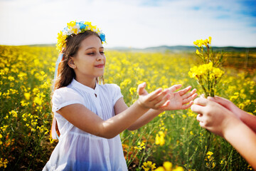 Girl in dress with Ukrainian wreath with flowers and ribbons takes from hands of unknown boy bouquet of rapeseed flowers