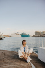 Happy young caucasian girl in sunglasses relaxing on weekend sitting pier by sea. Brown-haired woman with bob haircut wears casual clothes. Holiday life concept