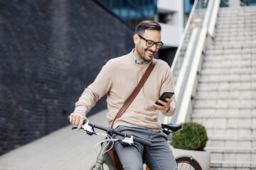 A casual businessman sits on a bike outside, scrolling on the phone and smiling at it.