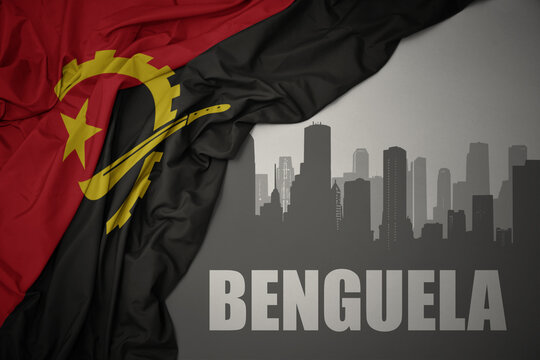 abstract silhouette of the city with text Benguela near waving colorful national flag of angola on a gray background.