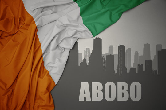 abstract silhouette of the city with text Abobo near waving colorful national flag of cote divoire on a gray background.