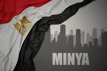 abstract silhouette of the city with text Minya near waving colorful national flag of egypt on a...