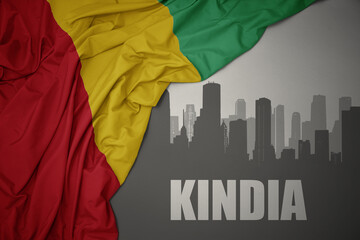 abstract silhouette of the city with text Kindia near waving colorful national flag of guinea on a...
