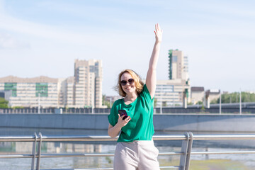 A happy young blonde student girl in sunglasses with a phone, waving her hand looking at the camera on the background of office buildings, river and blue sky. The girl greets friends. Date