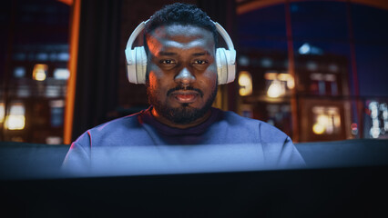 Obraz premium Young Handsome Black Man in Headphones Working from Home on Laptop Computer in Cozy Stylish Loft Apartment. Creative Male Smiling, Checking Social Media, Browsing Internet. Flat with Big Windows.