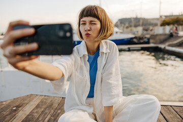 Cool young caucasian woman winks taking selfie on smartphone on background of yacht club. Brown-haired with bob haircut wears white shirt and pants. Summer vacation concept using