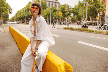 Modern young caucasian girl posing looking at camera outdoors. Brown-haired woman with bob haircut wears sunglasses, white shirt and pants. Lifestyle and fashion concept - obrazy, fototapety, plakaty