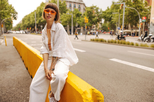 Modern young caucasian girl posing looking at camera outdoors. Brown-haired woman with bob haircut wears sunglasses, white shirt and pants. Lifestyle and fashion concept