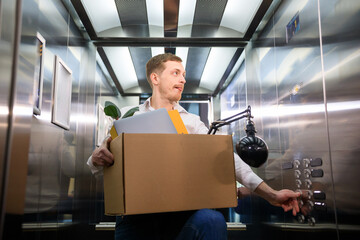 A young man holds a box with his belongings and tries to choose the right floor in the elevator