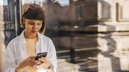 Attentive young caucasian woman uses smartphone standing outdoors at daytime. Girl with brown hair...