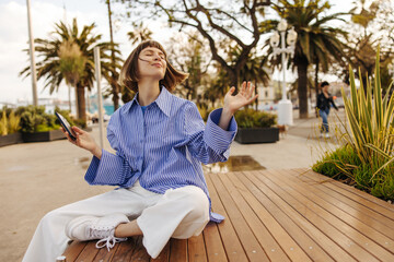 Satisfied young caucasian girl with phone closing her eyes is resting sitting on bench in springtime. Brown-haired woman with bob haircut wears casual clothes. Positive emotions concept