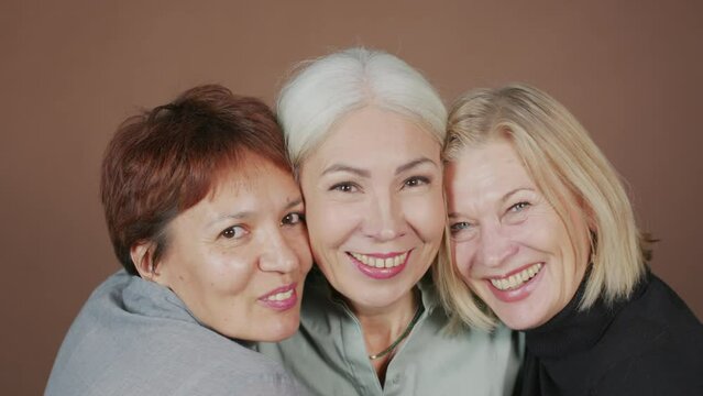 Medium closeup portrait of three smiling mature female best friends embracing each other while posing for camera on brown studio background