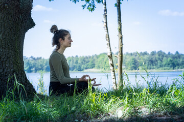 Young woman practicing yoga in nature. Sitting on green grass next to a tree and a forest lake. Communicate to exercise and maintain good health. High quality photo