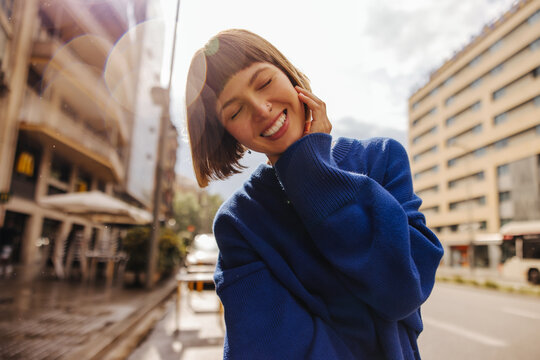Pretty young caucasian woman closing her eyes smiles teeth walking around city. Brown-haired with bob haircut wears blue sweatshirt. Positive emotions concept