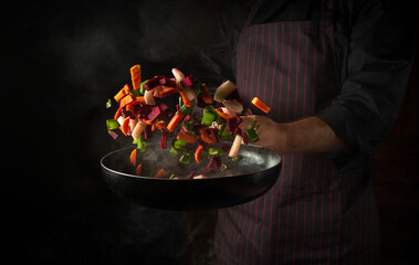 The hand of a professional chef throws pieces of vegetables into a hot frying pan with steam on a black background. The concept of cooking in the hotel. Free ad space. - obrazy, fototapety, plakaty