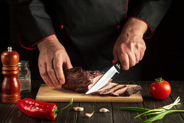 Experienced chef cuts the baked beef meat on a cutting board. The idea of preparing a delicious lunch