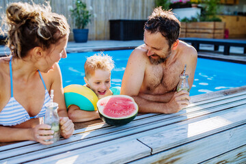Happy little boy with his parents in backyard swimming pool enjoying refreshments, drinks and...