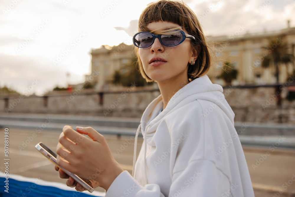 Wall mural Nice young caucasian woman with phone in her hands looks at camera walking outdoors. Brown-haired wears sunglasses, white hoodie. Lifestyle concept