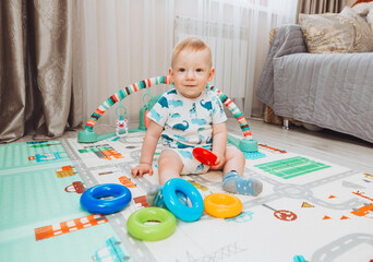 A cute baby is playing with a rainbow toy pyramid while sitting on a play mat in a sunny bedroom....