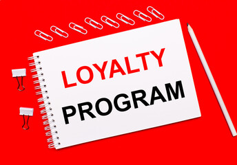 On a bright red background, a white pencil, white paper clips, and a white notebook with the text LOYALTY PROGRAM