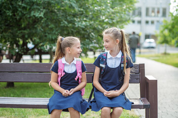 Fototapeta na wymiar Two children of schoolgirls sit on bench near school, have fun and communicate. Back to school