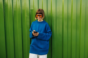 Positive young caucasian woman uses modern smartphone standing against green wall. Brown-haired...