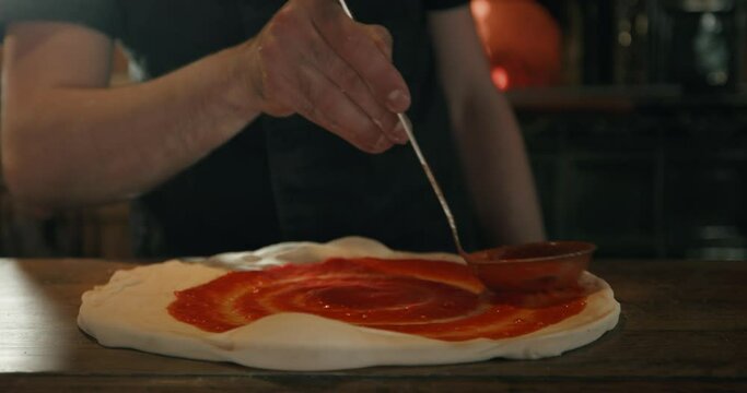 Pizza maker spreading tomato sauce on raw dough for pizza with ladle