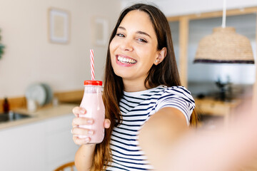 Healthy lifestyle concept with young attractive adult woman with homemade strawberry smoothy juice taking a selfie portrait with mobile phone in the kitchen at home