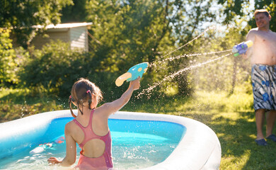 Little girl in swimwear, daughter dad, daddy, father swim, play water pistols, guns in children's inflatable pool in backyard of country house. Summer funny family outdoor games,recreation.Staycation