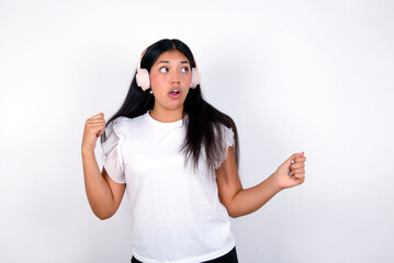 young brunette woman wearing white T-shirt standing against white background, dancing and listening music with headphones.