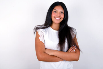 young brunette woman wearing white T-shirt standing against white background being happy smiling and crossed arms looking confident at the camera. Positive and confident person.